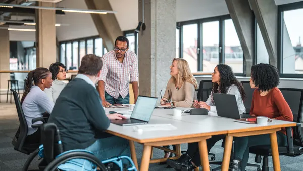 Indoor shot of a big group of multi ethnic people and one disabled man having a work meeting during the day. They are all seated and the male boss is standing.