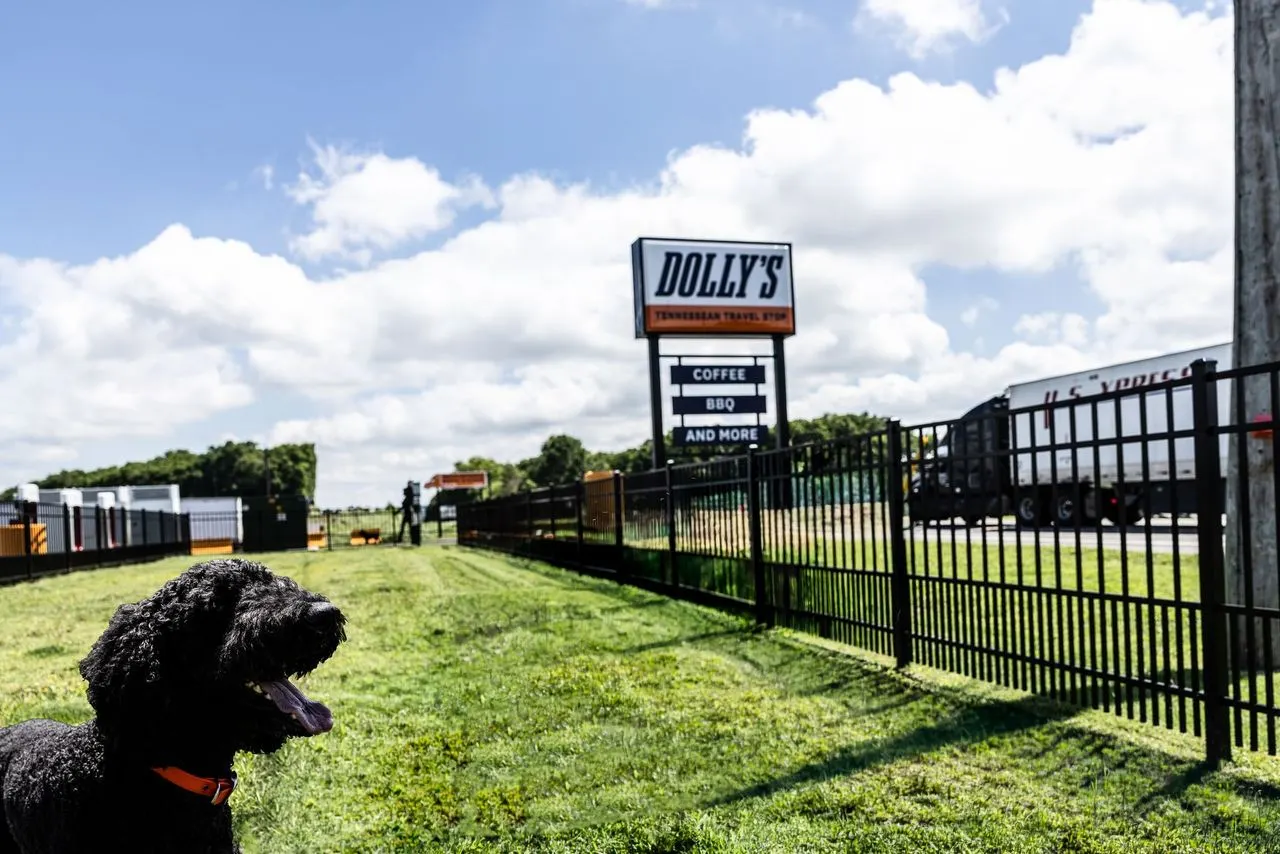 A photo of a dog at a dog park. The space is fenced in with a metal fence and a sign in the background says Dolly's Tennessean Truck Stop.