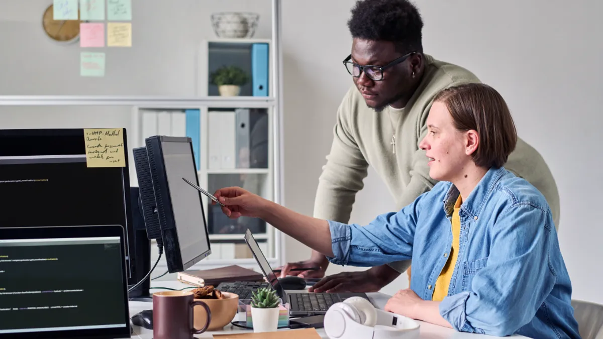 Young woman pointing at computer monitor and discussing new software together with her colleague during teamwork at office