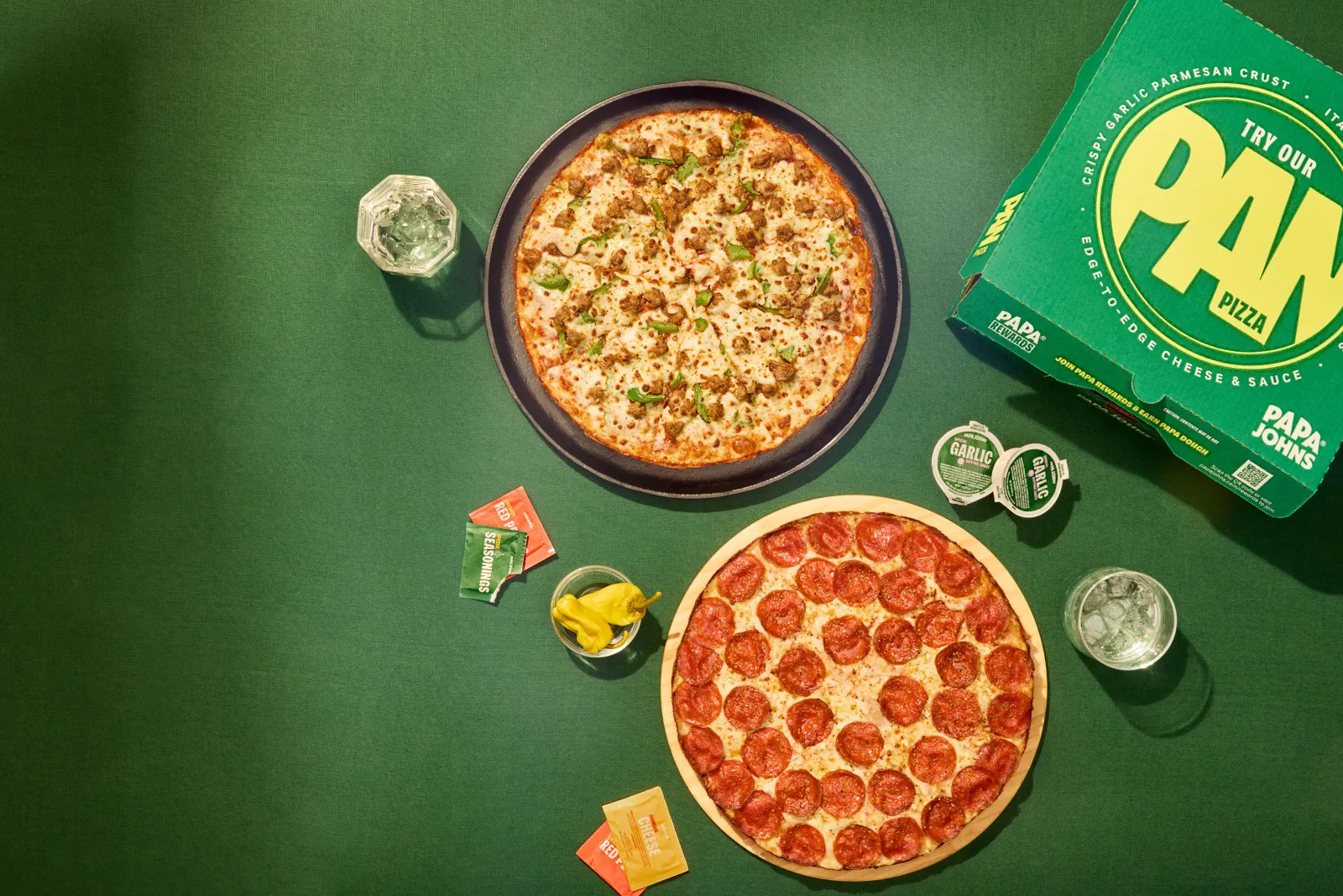 An image of two pan pizzas in front of a green background. On the right is a green pizza box.