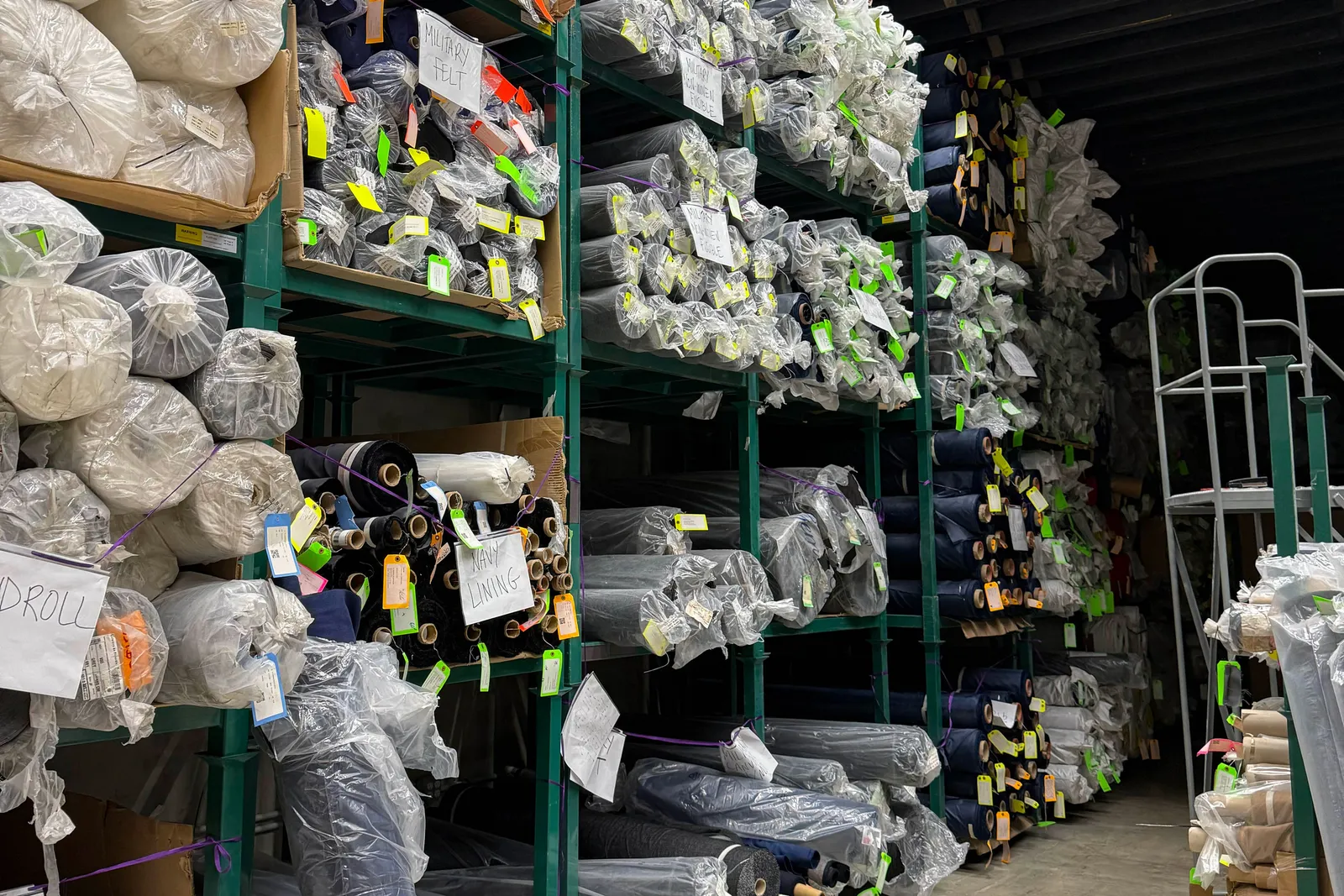 Rolls of stacked fabric, some in plastic sheeting, sit in a warehouse on multiple shelves.