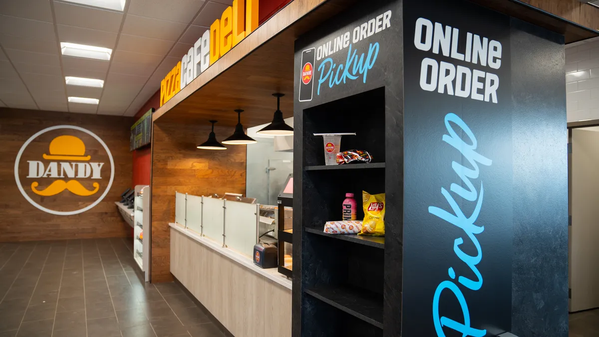 A photo of the interior of a convenience store, showing a pickup area and ordering area for food. A sign on the wall says "Dandy", a sign over the food ordering areas says "pizza deli cafe" and the pickup area says "online order pickup"