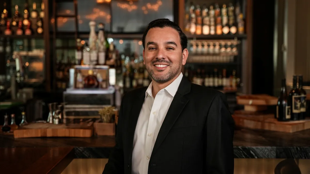 A smiling white man in a dark suit with no tie in front of a bar.