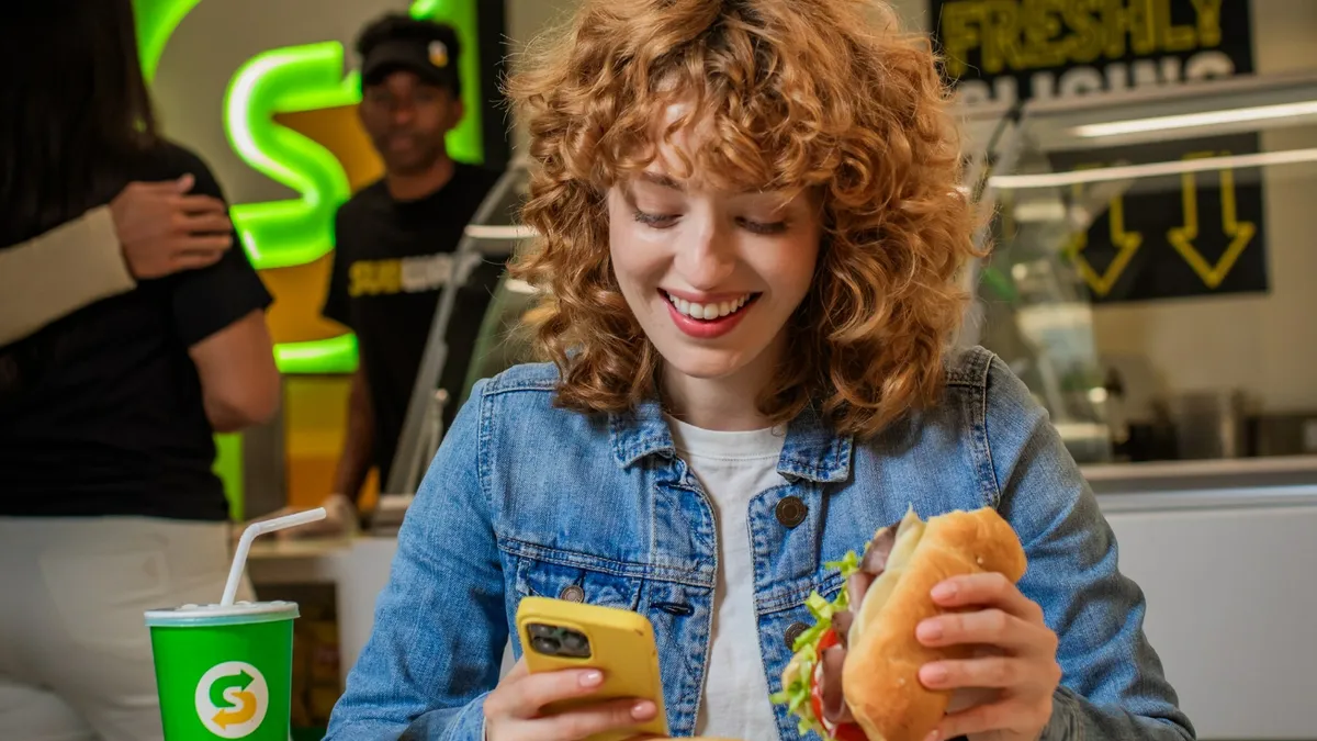 A woman grins at her phone while eating a Subway sandwich in a promotion image for the chain's MVP Rewards.