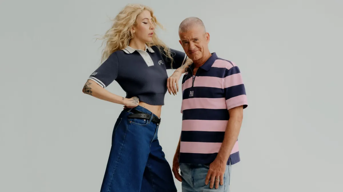 A woman stands on a box next to a man and leans on his shoulder. Both wear casual attire.
