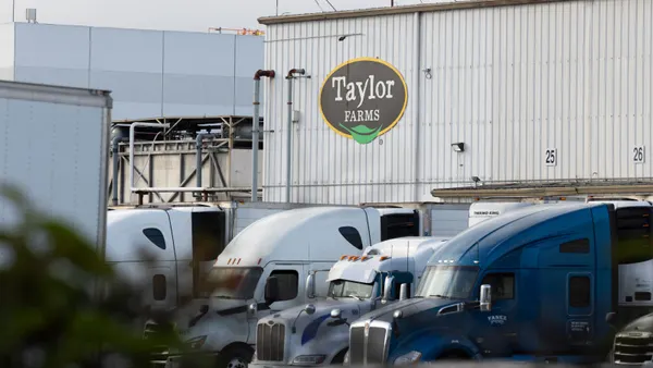 Trucks sit parked outside a logistics facility. The building has the "Taylor Farms" logo printed on its side.
