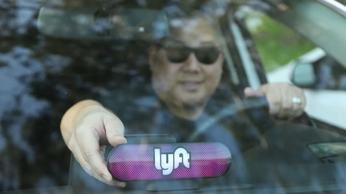 A driver seen through the windshield of a car. The driver is adjusting a device on the dashboard that reads "lyft."