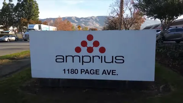 A white sign with the black and orange Amprius logo, with mountains and a parking lot in the background.