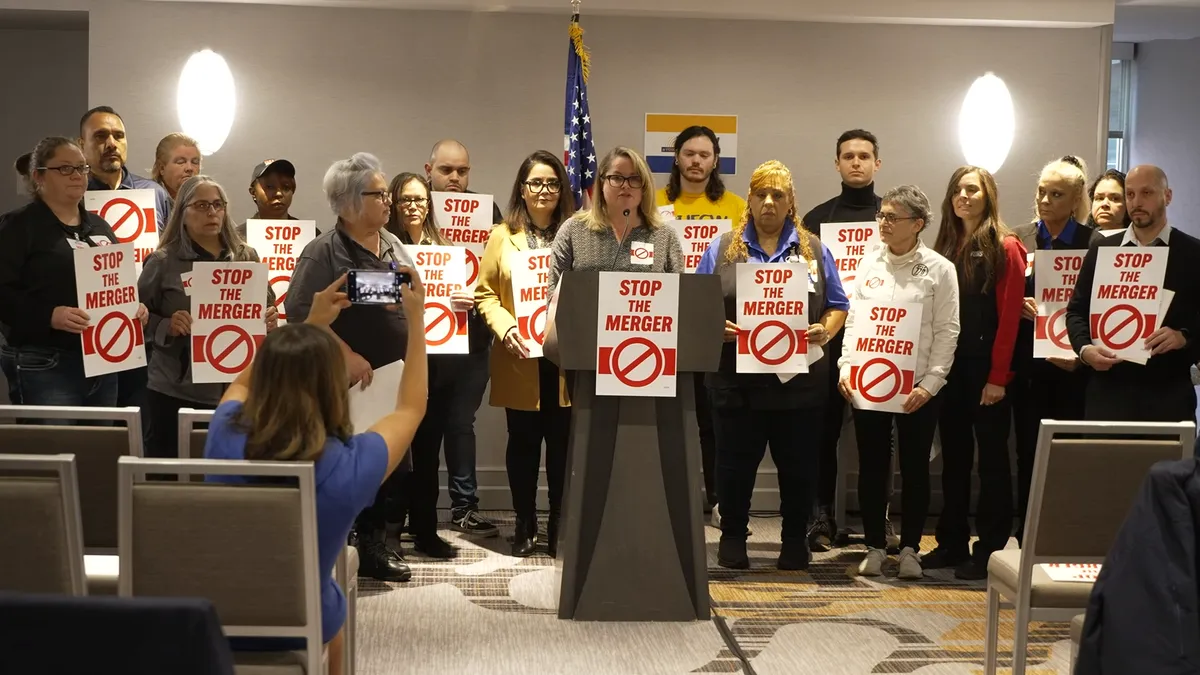 More than a dozen people with "Stop the merger" signs standing behind a podium