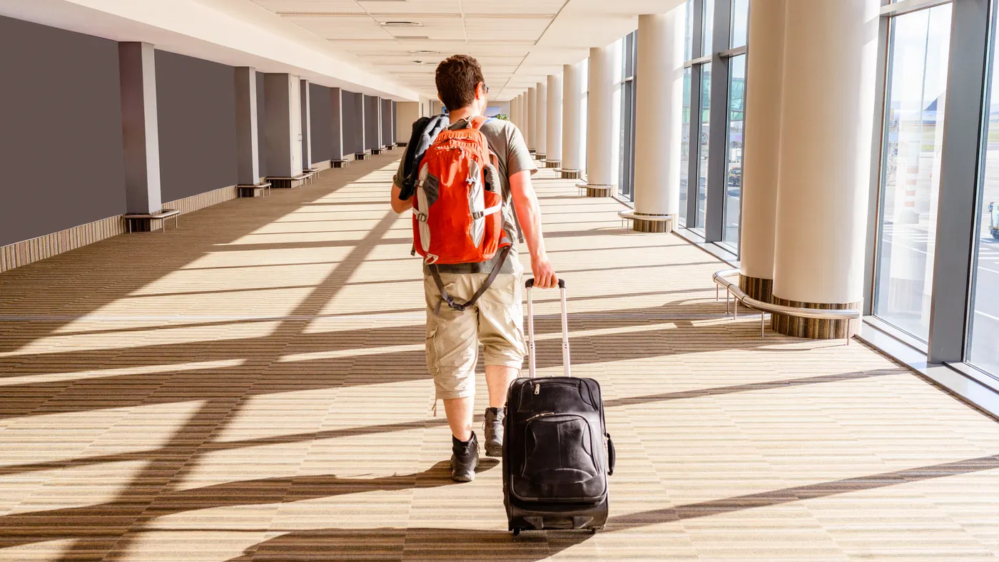 A young person wearing a backpack drags a rolling suitcase behind them as they walk through an airport.