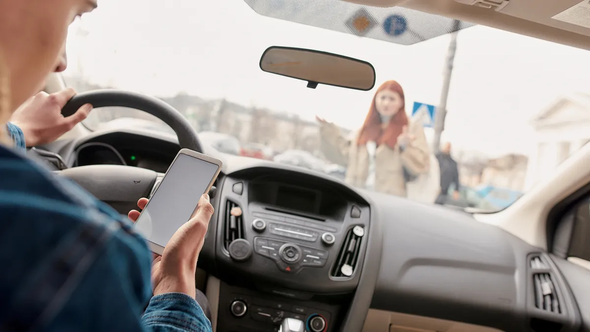 A distracted driver looks at their smartphone while someone crosses the street in front of their vehicle.