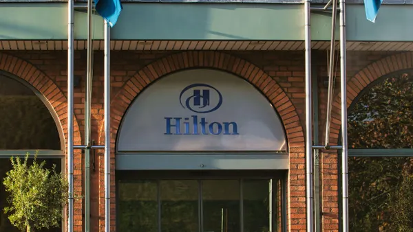 Front view of a brick building featuring a large arched entrance topped with a silver plaque displaying the blue Hilton logo. Two flags are mounted above the entrance, and the facade includes tall windows and decorative stonework.