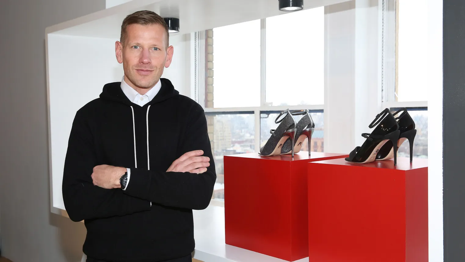 A man stands with his arms crossed next to two pairs of high-heeled shoes.