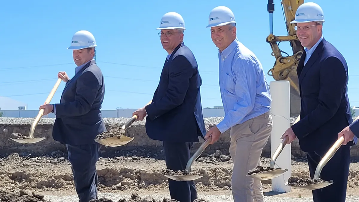 Four people wearing hardhats use shovels to throw dirt at a groundbreaking ceremony.