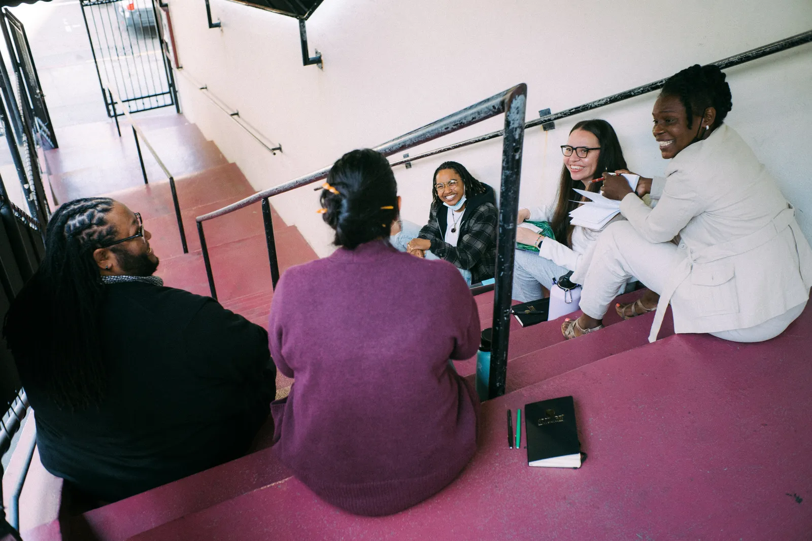 A group of adults are sitting on a staircase on different steps with two having their backs to the camera and three facing the camera.