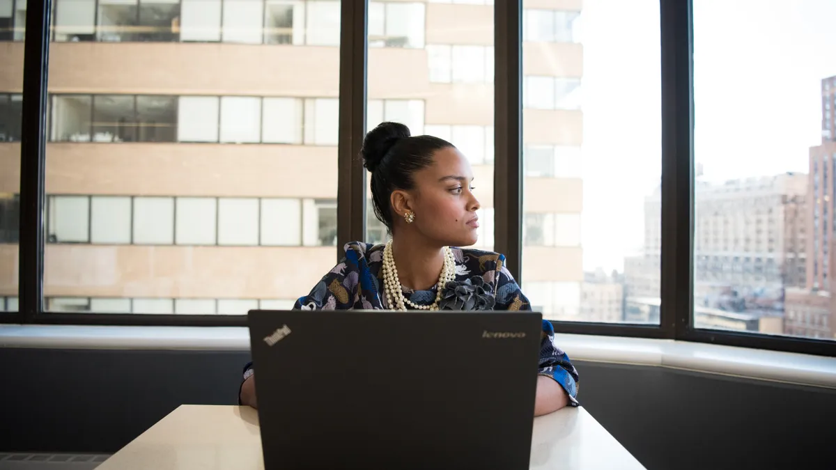 Professionally-dressed woman sits in front of black laptop in office.