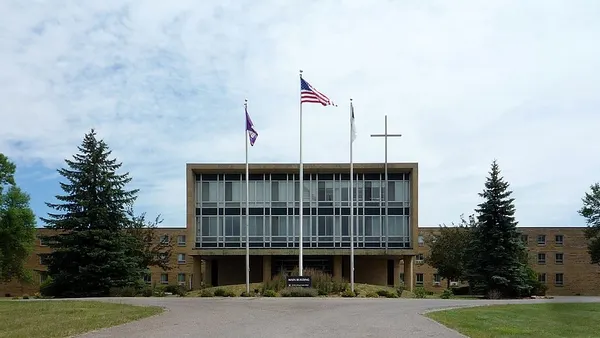 A campus building sits in the middle ground, behind a large cross and a flag pole with the U.S. flag.