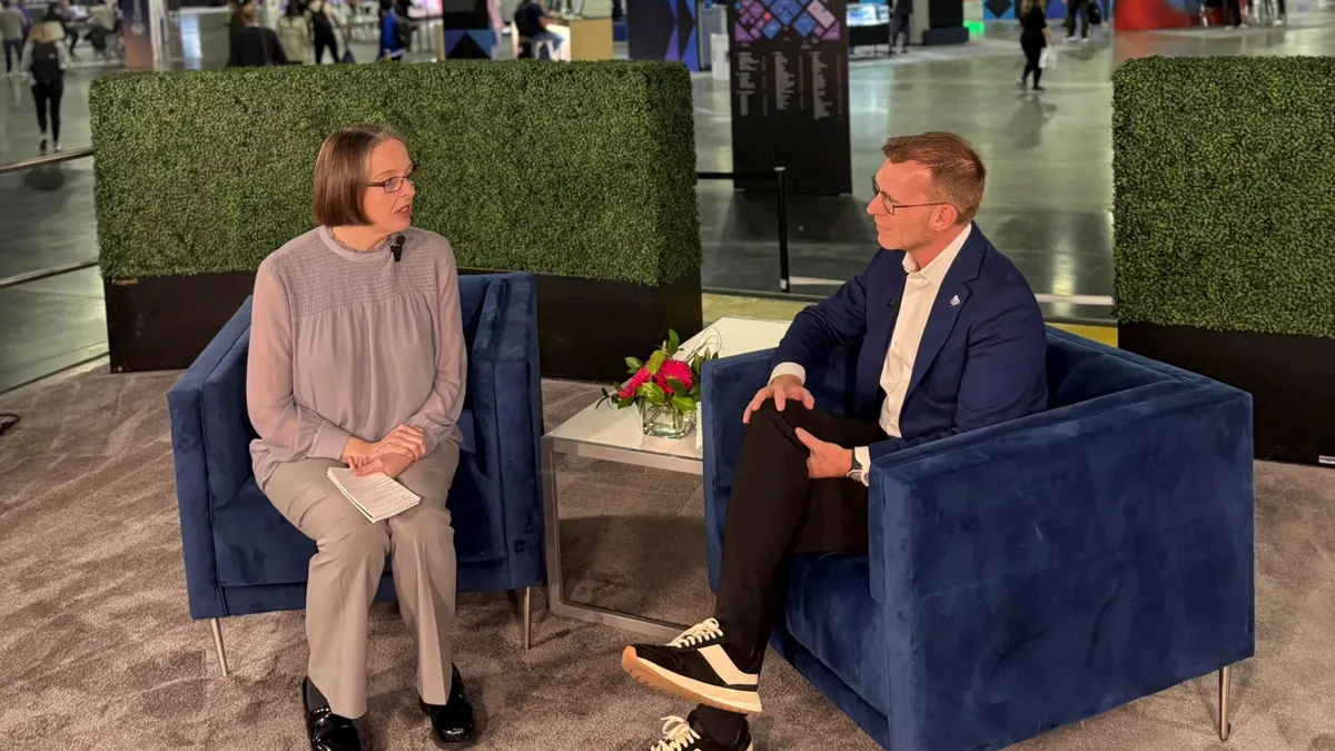 TechTarget lead editor Beth Pariseau and IBM outbound CTO for automation Tyler Lynch are seated in blue chairs. Pariseau is on the left wearing gray pants, black shoes, lavender blouse; Lynch on the right is wearing black and white sneakers, black pants, a white shirt and a blue jacket. Behind them is a hedge divider, and the IBM TechXChange 2026 conference expo floor is beyond that.