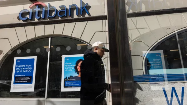 A Citi bank branch is shown, as a person walks by. Window signs advertise the bank's offerings.