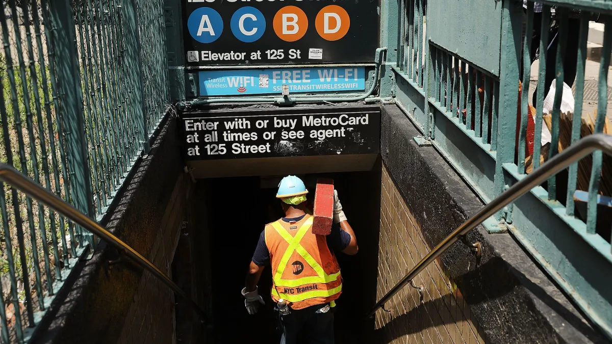 A man wearing a hardhat and safety vest climbs down the stairs to a subway station.