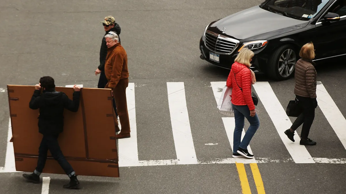 Pedestrians walking though an intersection in New York City in front of a black Mercedes-Benz sedan.