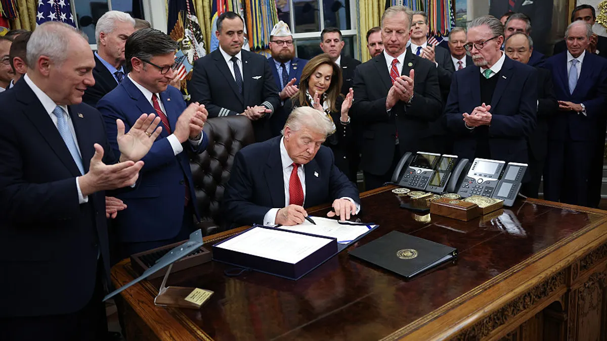 A person is sitting at a desk signing a piece of paper. Other people are standing gathered around the desk and clapping
