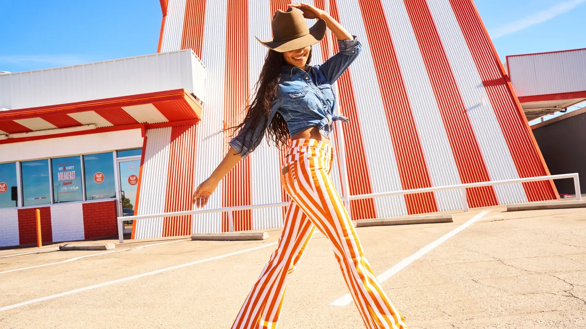 A person in a blue denim top, cowboy hat and orange and white striped flare jeans smiles as she hold on to the top of her hat and walks past an orange and white Whataburger location.