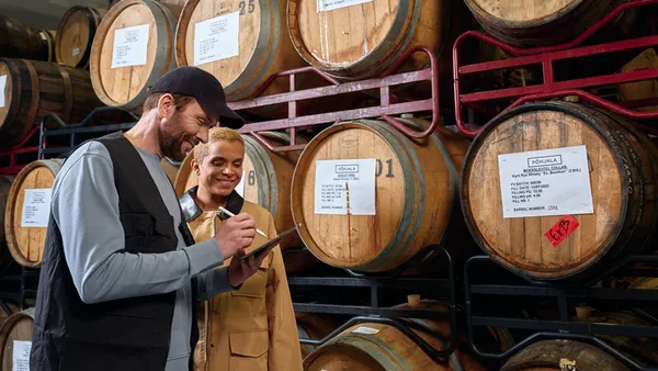 Two people in a storage area with wooden barrels. One is writing on a clipboard, both smiling. The scene conveys a collaborative and happy atmosphere.