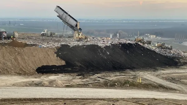 A truck dumps waste onto the open face of a landfill at dawn. Other trucks, workers, and landfill gas wells are visible.