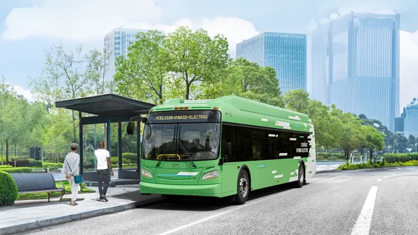 A green bus at a bus stop with two people on the sidewalk and tall buildings in the background.
