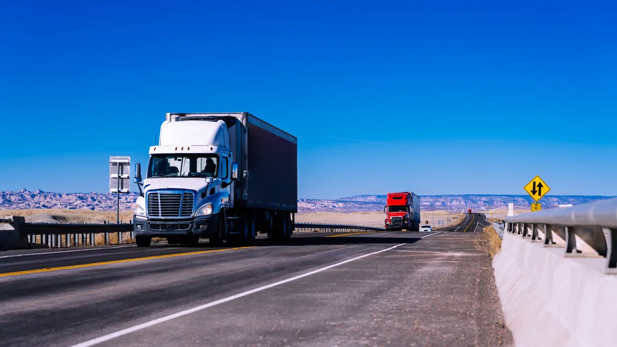 Tractor-trailers on a desert highway with clear blue sky and rocky landscape surrounding the roadway. A yellow highway sign indicates a curve ahead.