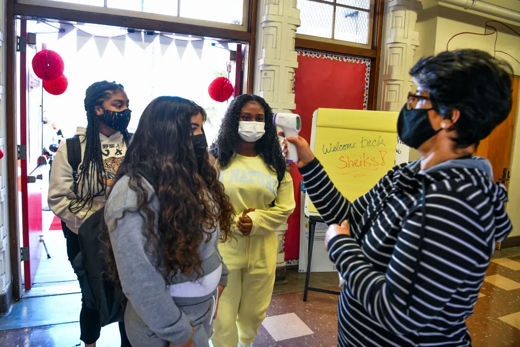 Three students stand in the entrance of a school. A school staffer is holding a thermometer to take a student's temperature.