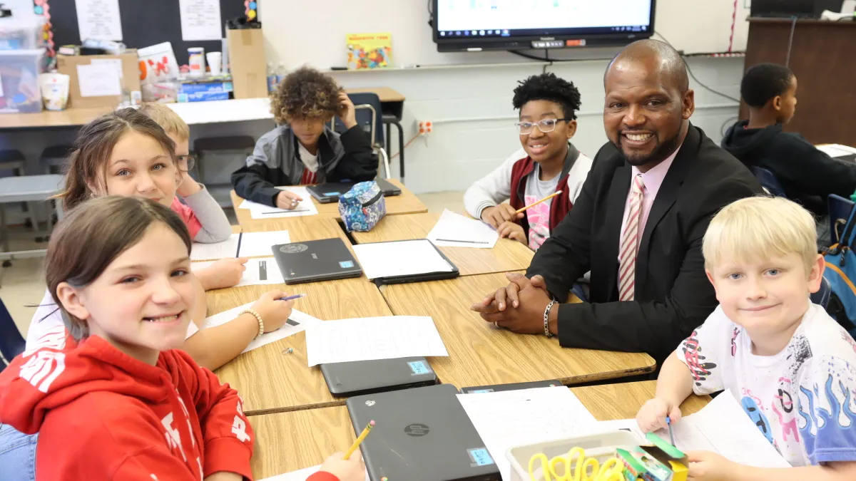 An adult sits at a table with children. Paper, laptops and other materials are on the table.