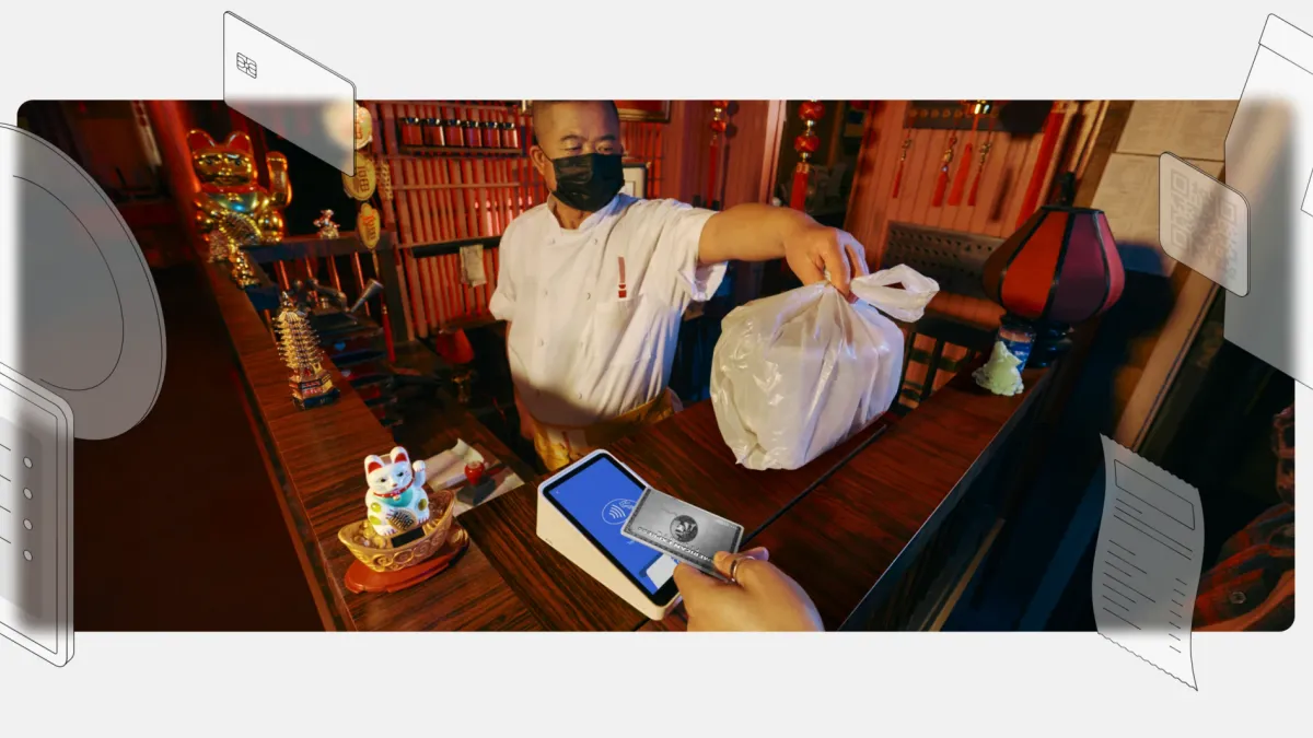 A worker at a restaurant with a face mask on hands over a bagged order while an offscreen person taps their credit card on a Square card reader.