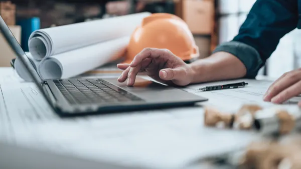 A person works on a laptop in the foreground of an artistically minded shot. In the background are blueprints and a yellow hardhat.