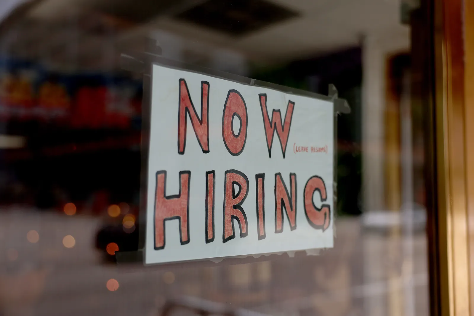A handwritten "now hiring" sign hangs in a shop window.