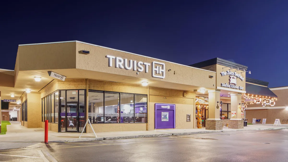 A Truist bank branch located in a shopping center at night, with illuminated signage and a purple ATM on the exterior wall. The building has large windows showing the interior, and adjacent businesses including a seafood restaurant named "The Juicy Seafood" and a dentist office are also visible.