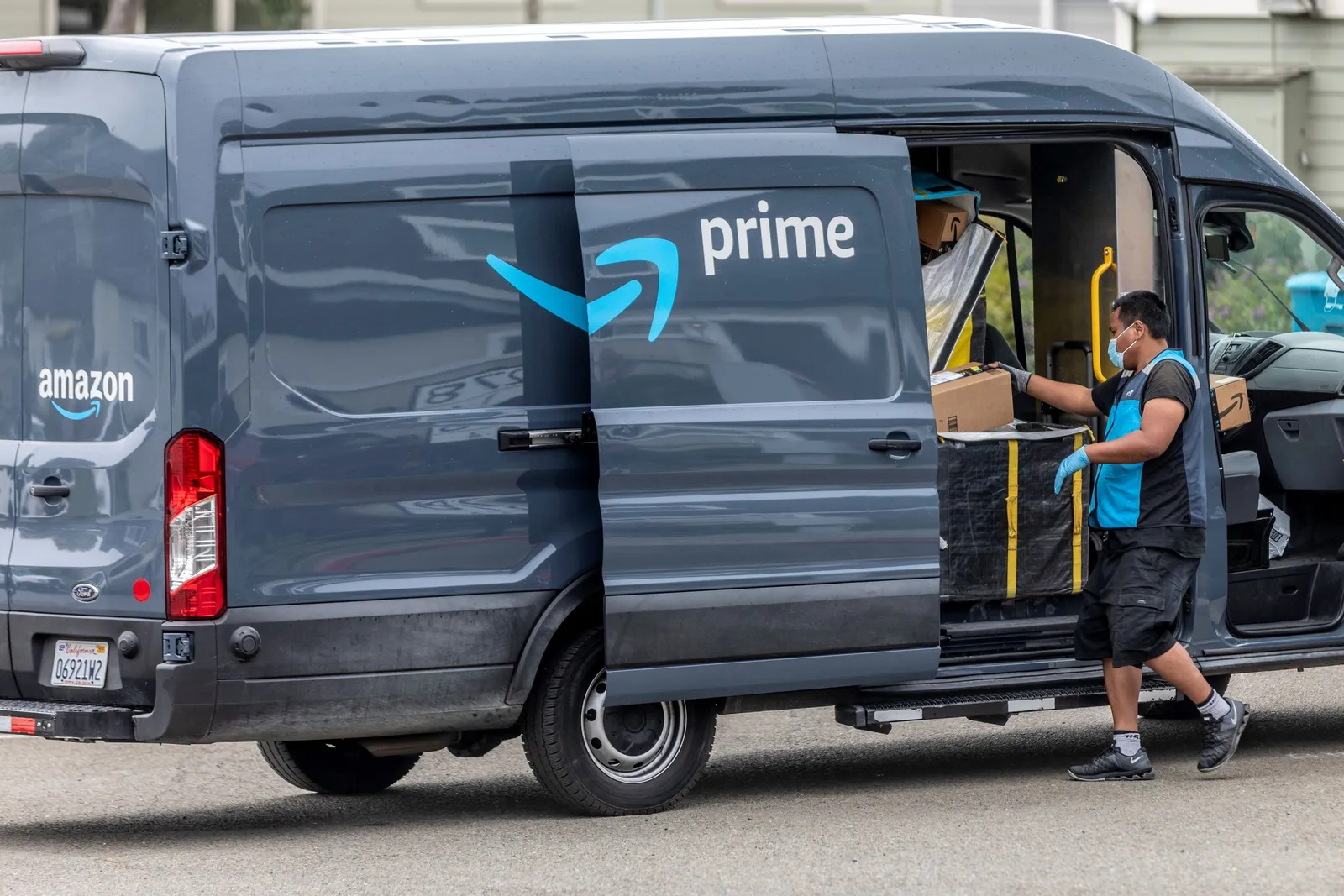 A worker wearing a mask and gloves stands outside of an Amazon Prime van containing packages.