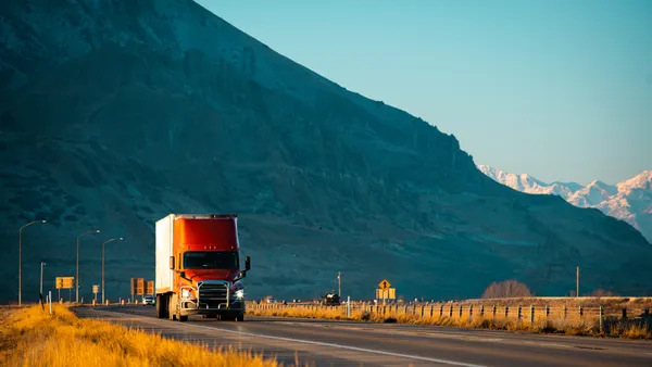 A red semi-truck with lights on and a white trailer attached drives along a winding Utah highway with towering, rugged mountains dusted with snow in the background.