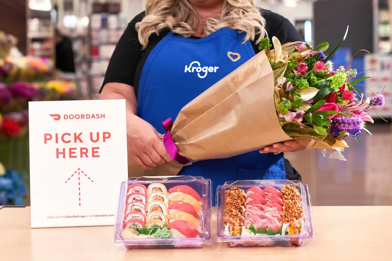 Image of a Kroger worker holding flowers in front of sushi packages by a &quot;DoorDash pick up here&quot; sign