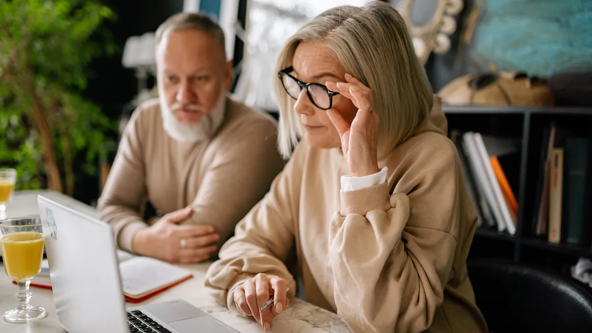 Older couple sits at a table, with one person sitting at the laptop