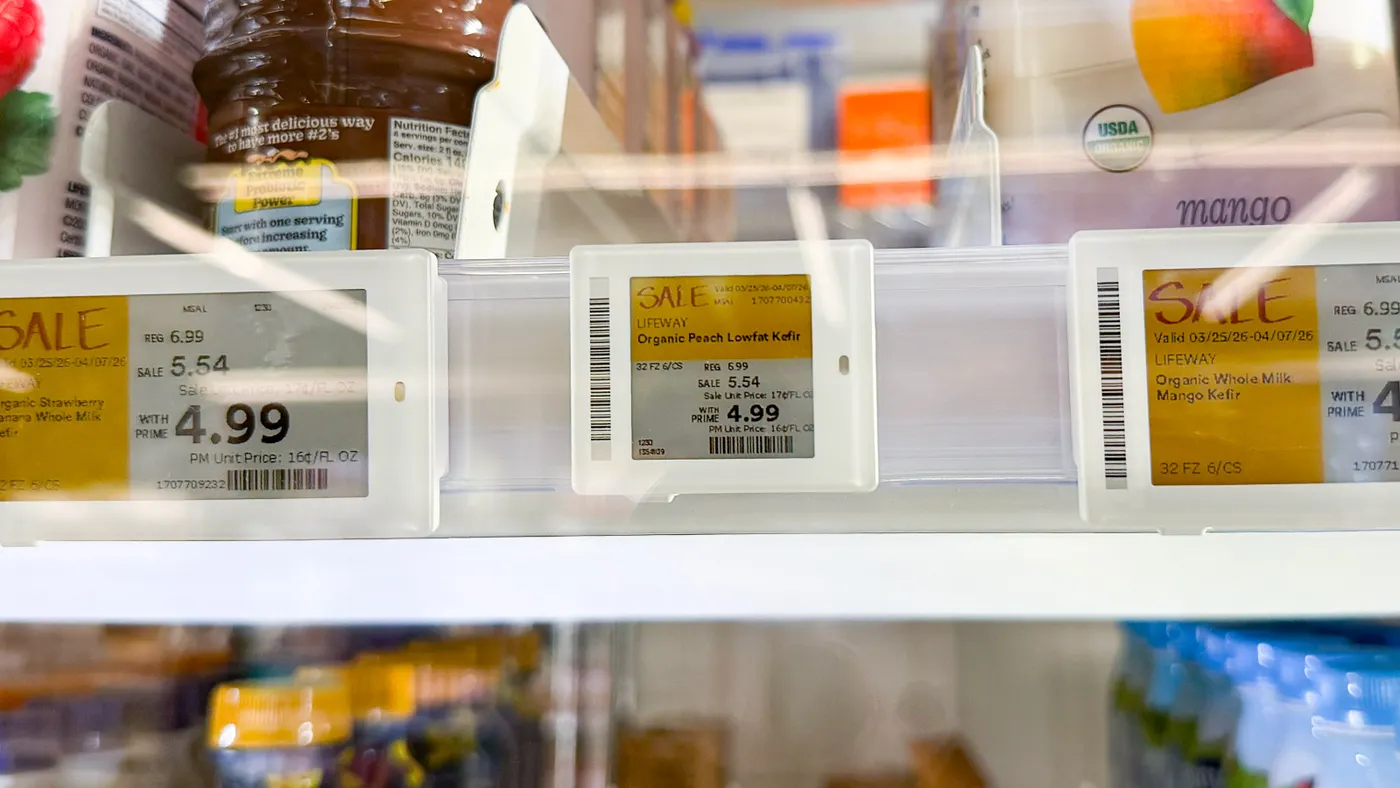 Close-up of electronic shelf labels in a refrigerated case displaying “SALE” prices for Lifeway kefir, including “Organic Peach Lowfat Kefir,” with pricing shown as “REG 6.99,” “SALE 5.54,” and “WITH PRIME 4.99,” and blurred dairy containers behind.
