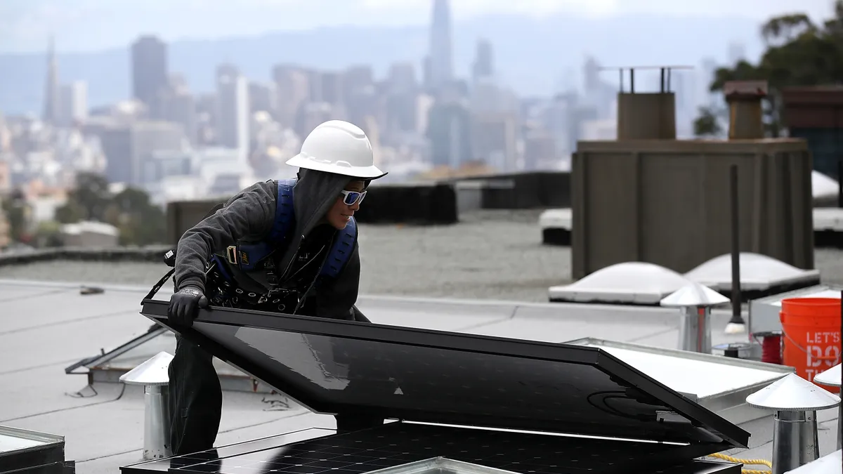 A person in a hard hat lifts a solar panel on a building in front of the city skyline.