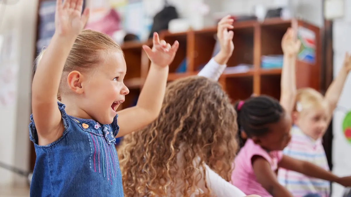 A handful of young children are seated on the floor in a row. They are looking forward and some have their arms raised.