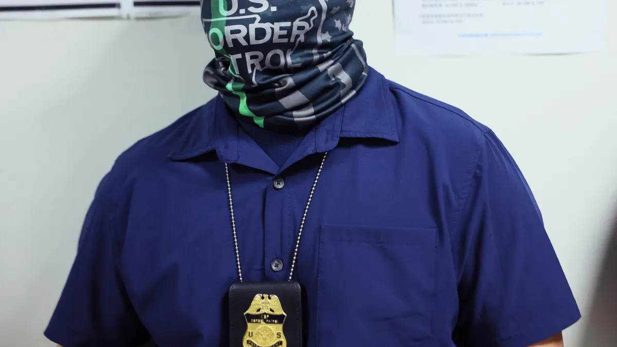Person with face wrapped in scarf that says "US Border Patrol" and badge on a chain around their neck.