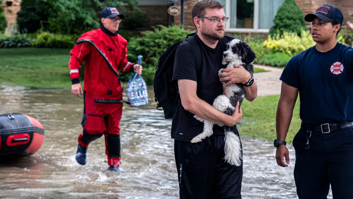 A person carrying a wet dog wades through a flooded street while two people look on.