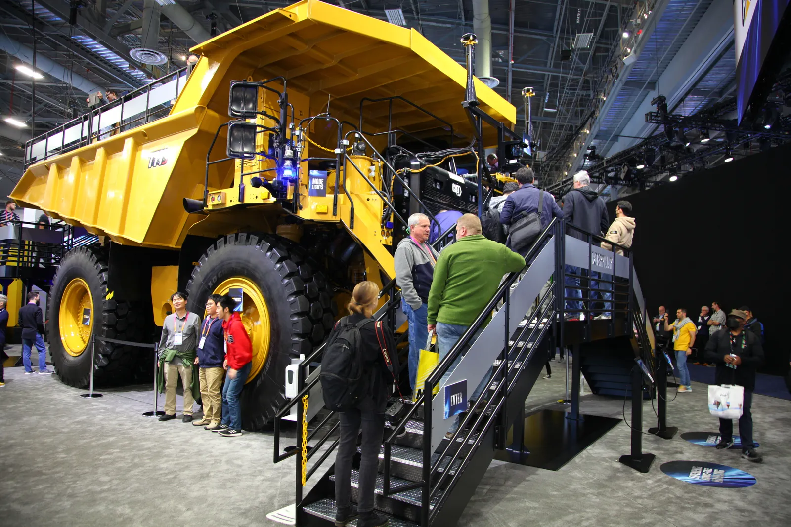 A group of people on a convention center floor stand in front of a heavy machinery truck that was made by Caterpillar.