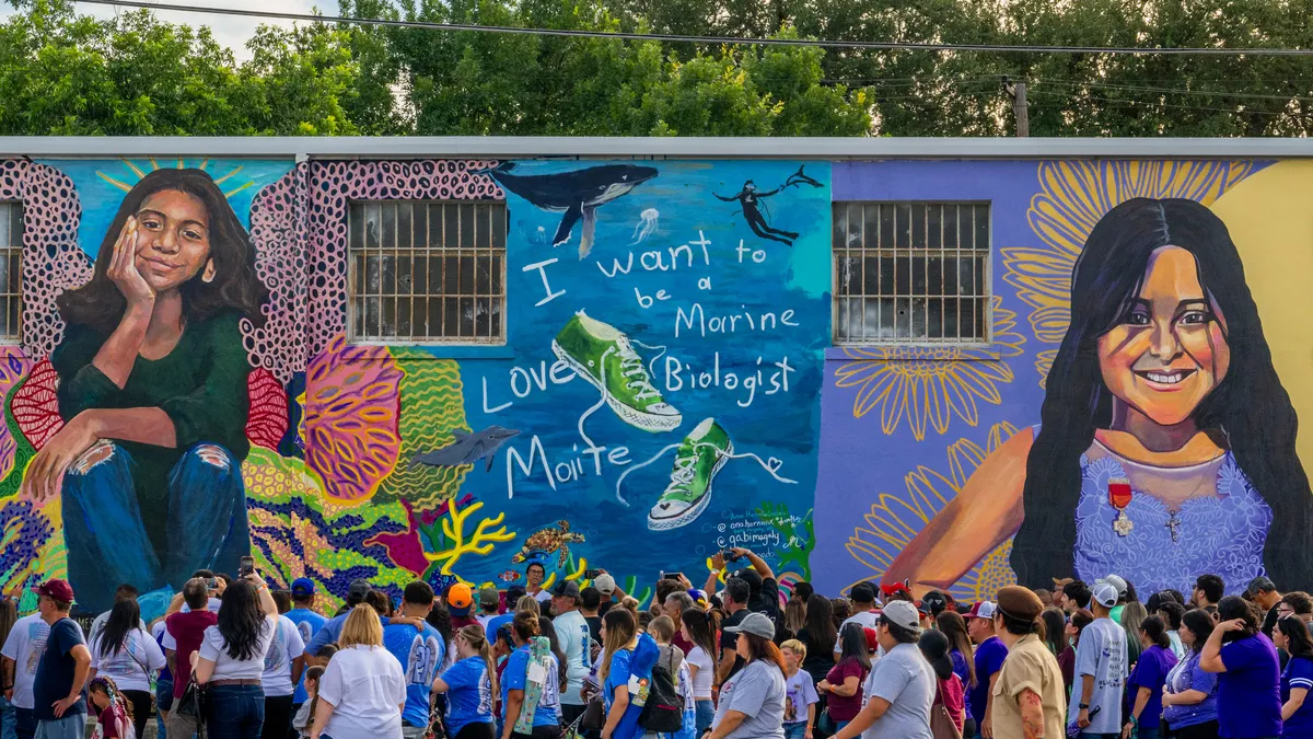 A crowd of mourners stand in front of a mural for Uvalde victims