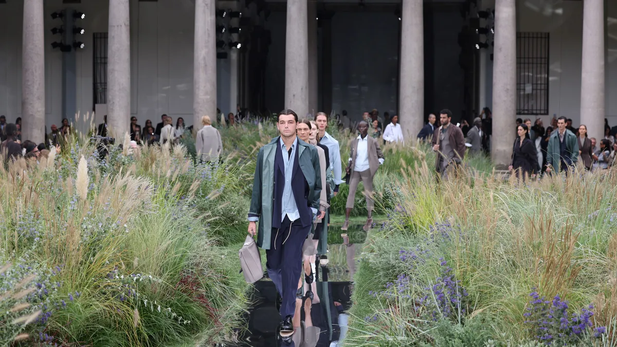 Models in suits walk between patches of tall grass on a fashion runway.