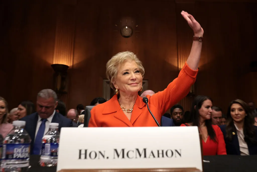 A person is seated as a desk. They are waving their left hand. A name plate sits in front of them and people are seated in row in the background.